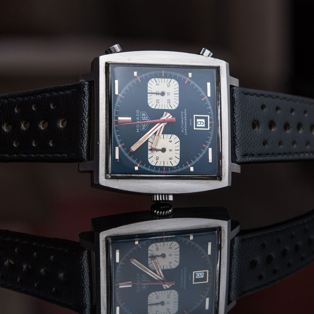Die Uhrenikone TAG Heuer Monaco mit links platzierter Krone liegt auf einem gläsernen Tisch.